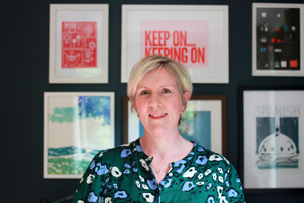 Harriet Mason in her office. Blonde hair, green floral patterned dress against a navy wall and brightly coloured pictures