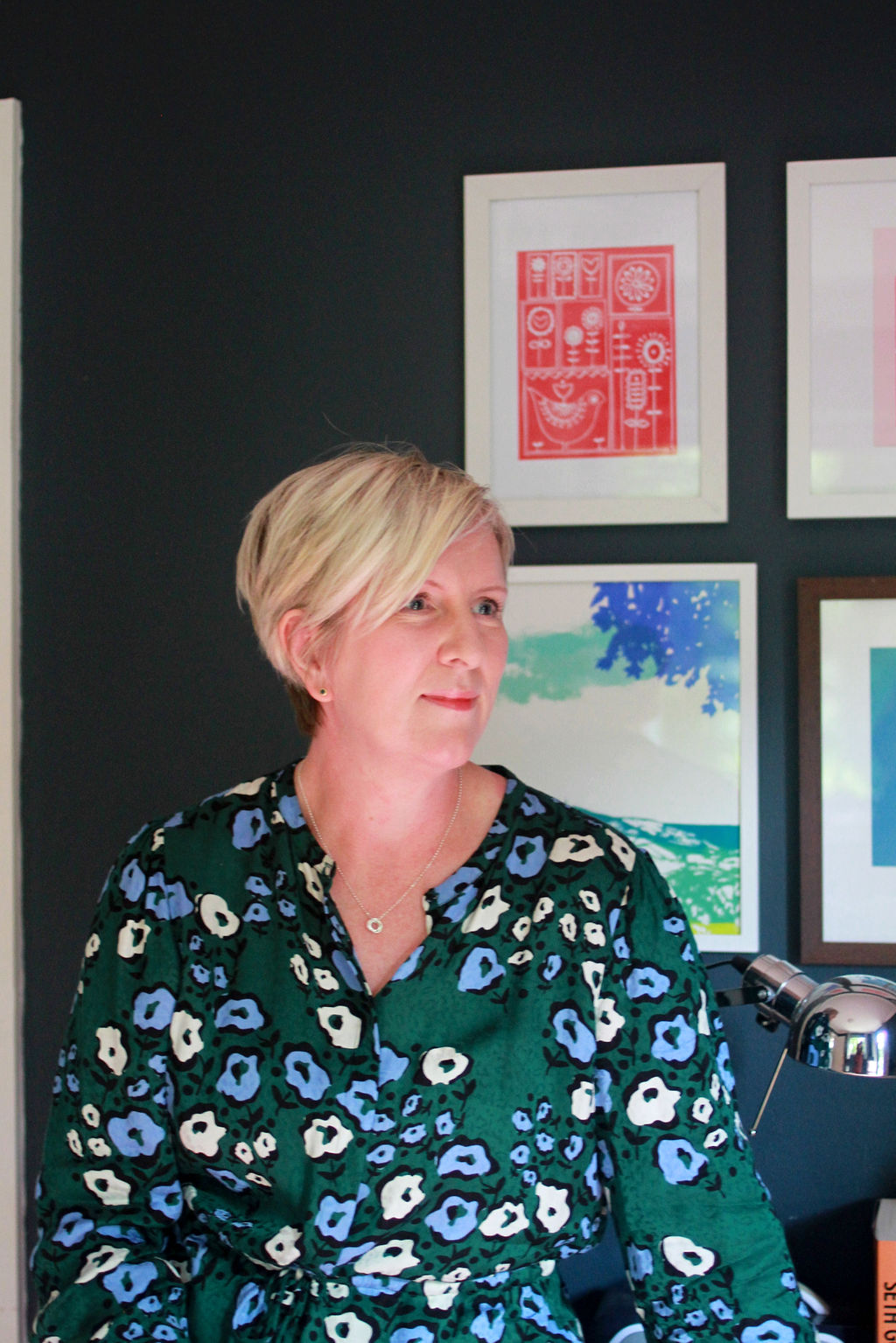 Harriet Mason in her office. Blonde hair, green floral patterned dress against a navy wall and brightly coloured pictures