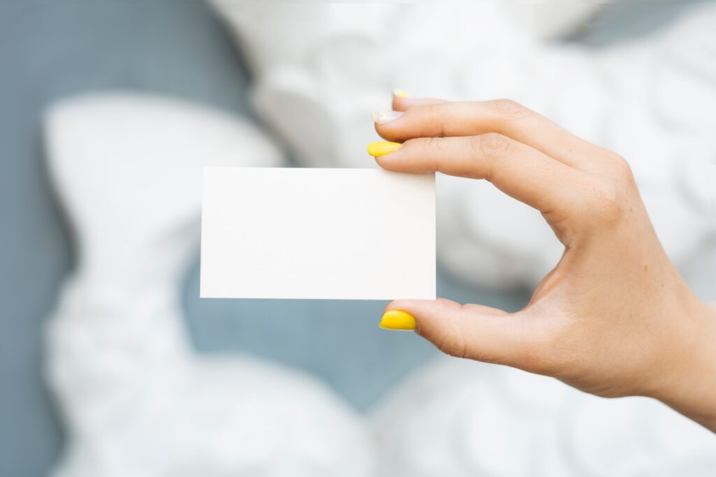 A woman's hand with bright yellow nail varnish holding a blank index card against a blurry white and blue background