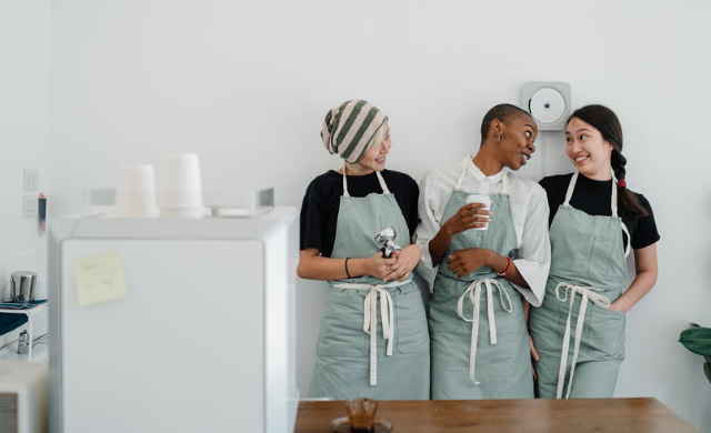 Three young women in their twenties leaning against a white wall in a large kitchen. They're wearing the same pale green apron, looking at each other smiling and laughing