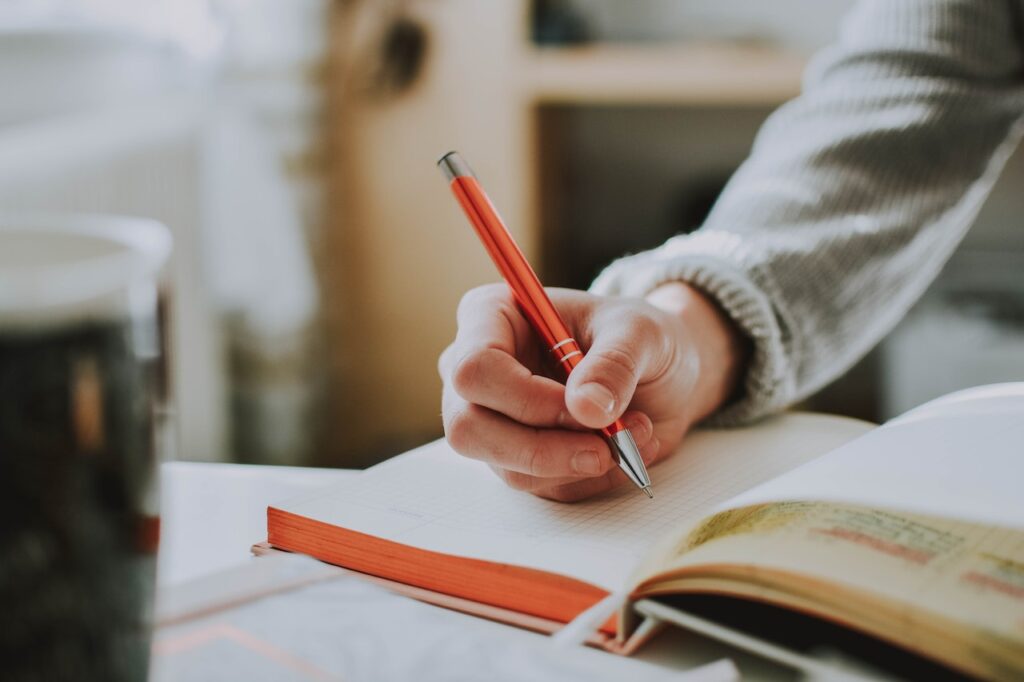 Image is a close up of an open notebook, with someone's arm hovering above the open page, holding an orange pen as if about to write something down