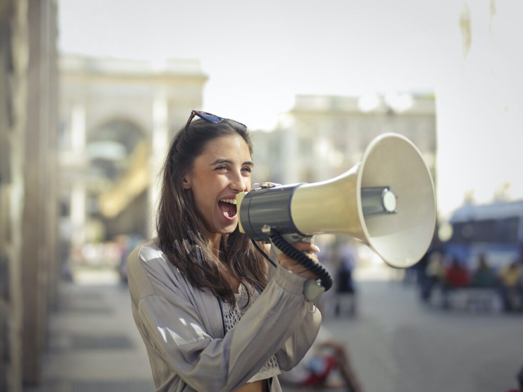 Young woman with long back hair and sunglasses on her head holding a large loud hailer. Her mouth is open and she's smiling, as if she's using the loud hailer to make herself heard.