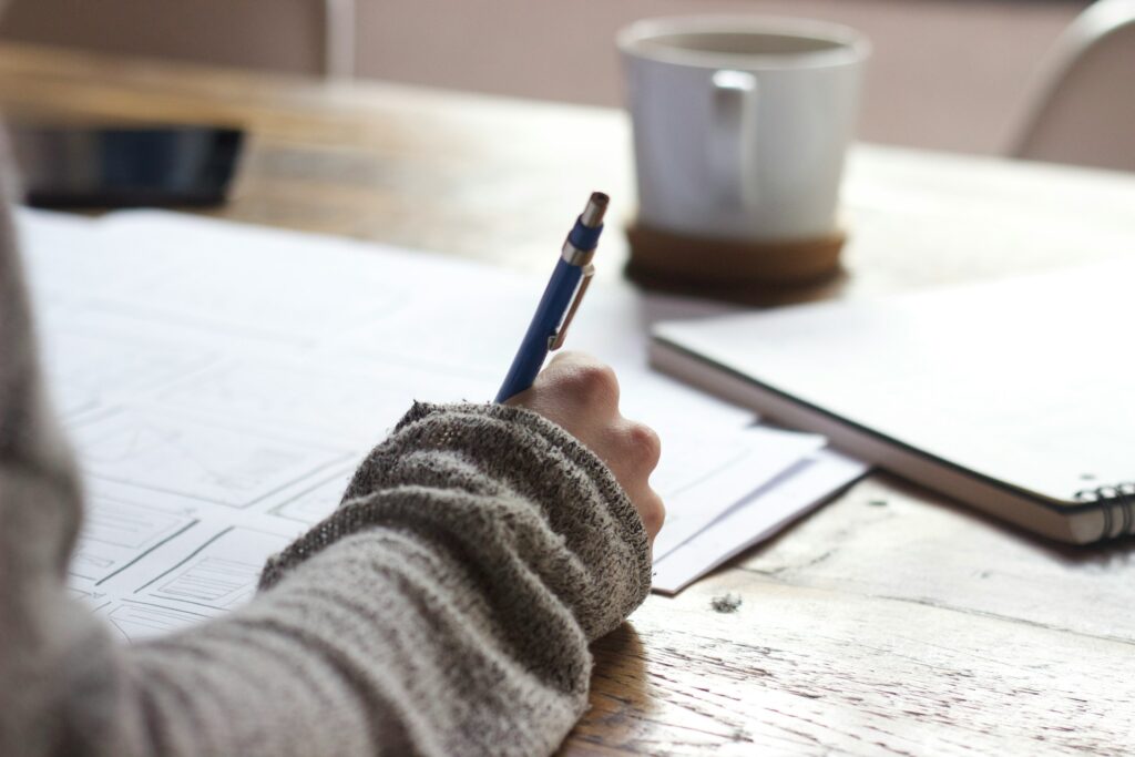 Just seen persons arm writing with a pen on a white paged open notebook at a wooden desk with a white cup of coffee or maybe tea