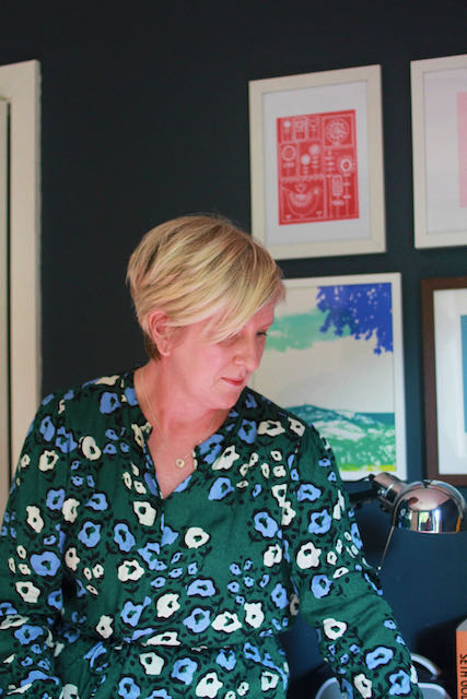 Harriet Mason in her office. Blonde hair, green floral patterned dress against a navy wall and brightly coloured pictures