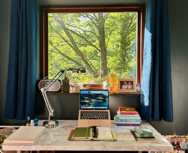 Harriet's desk under a square window looking out at trees, covered in green leaves. The desk is covered with notebooks, a laptop, anglepoise lamp and piles of books.