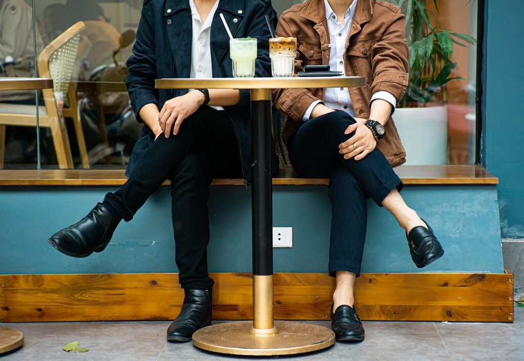 Two people sitting side by side at a round bistro style cafe table. We can't see their heads, only from the shoulders down. Both cross their leg away from each other, their hands on their laps, two coffees on the table