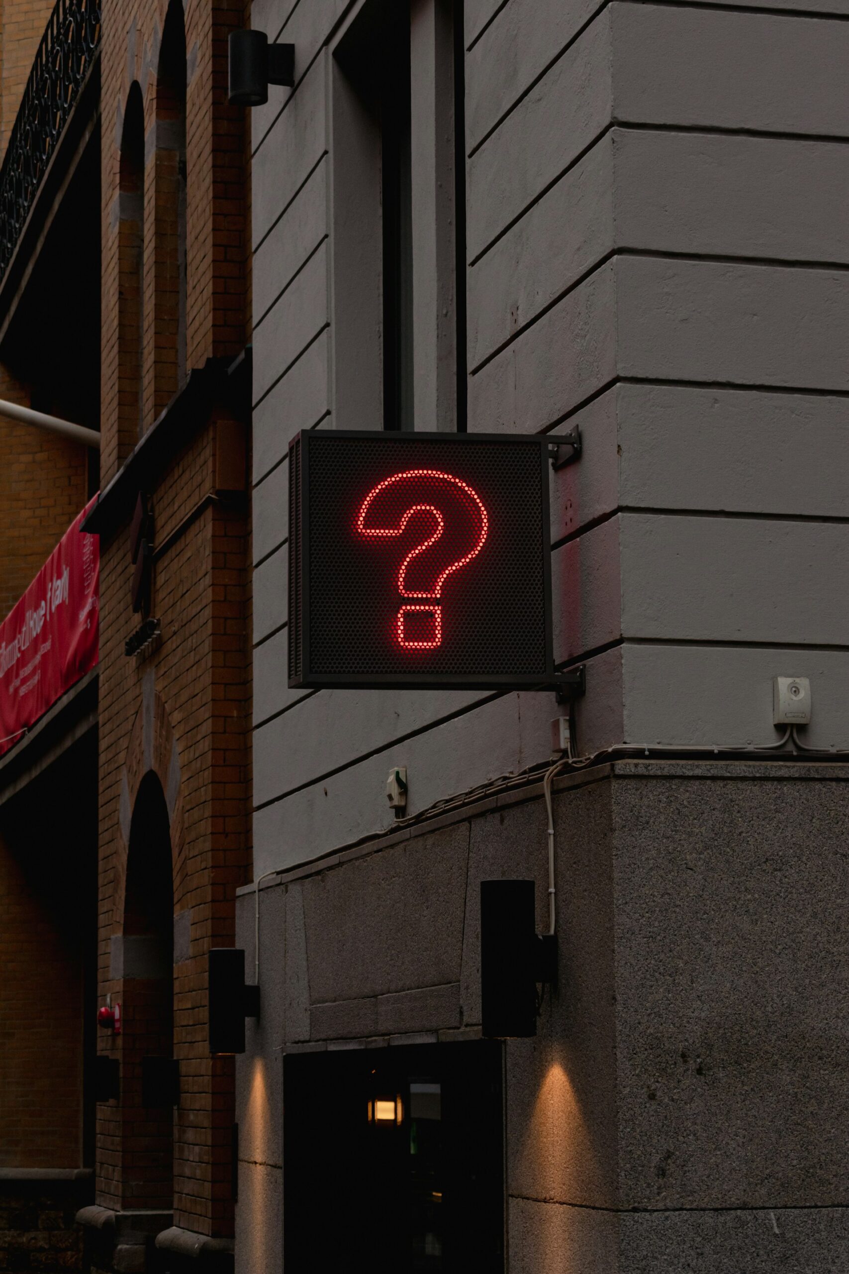 Image is of the corner of an urban building with a black sign with a red illuminated question mark on it.