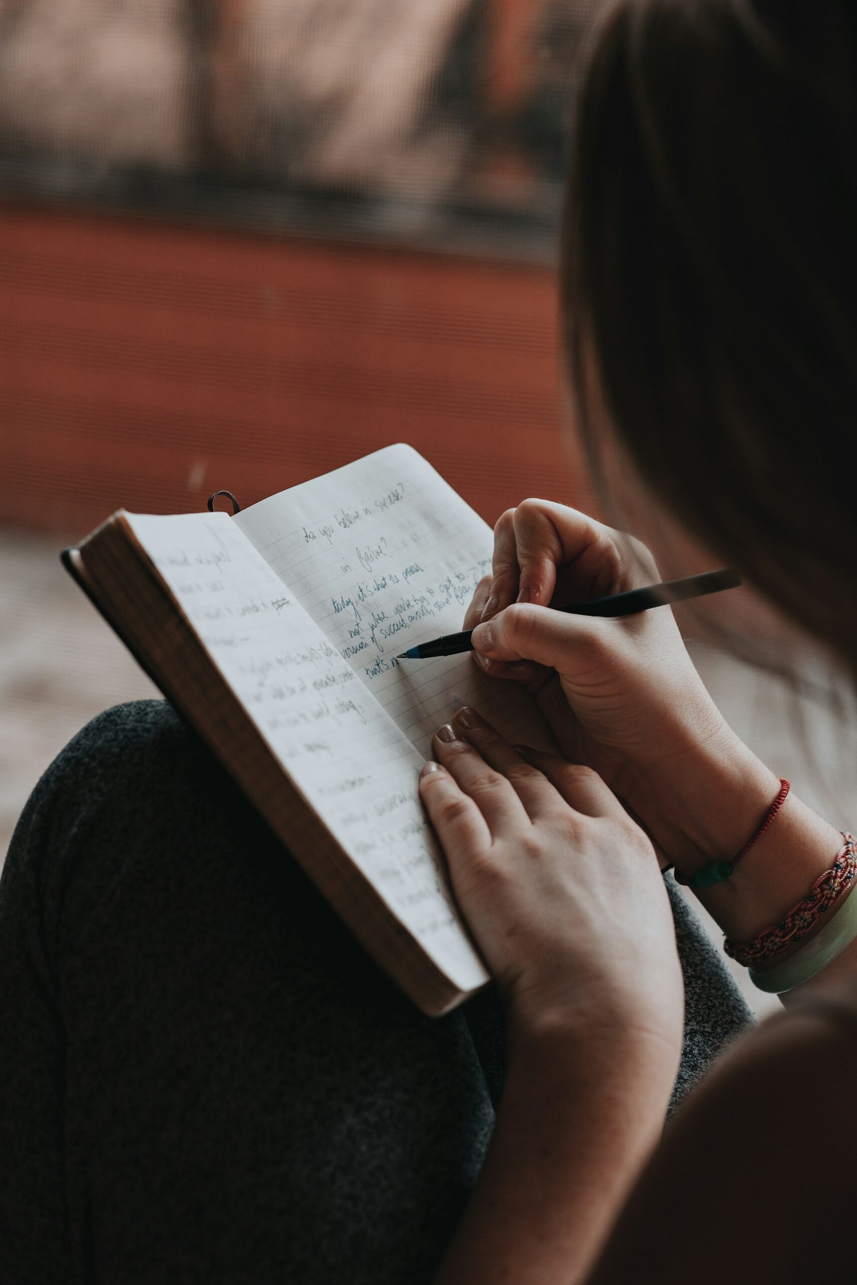 Image is of a woman sitting, a notebook open and resting on her knees. She's writing with a pen in her right hand. We can't see her face, her long brown hair is shielding her face and her left hand is holding the notebook open.